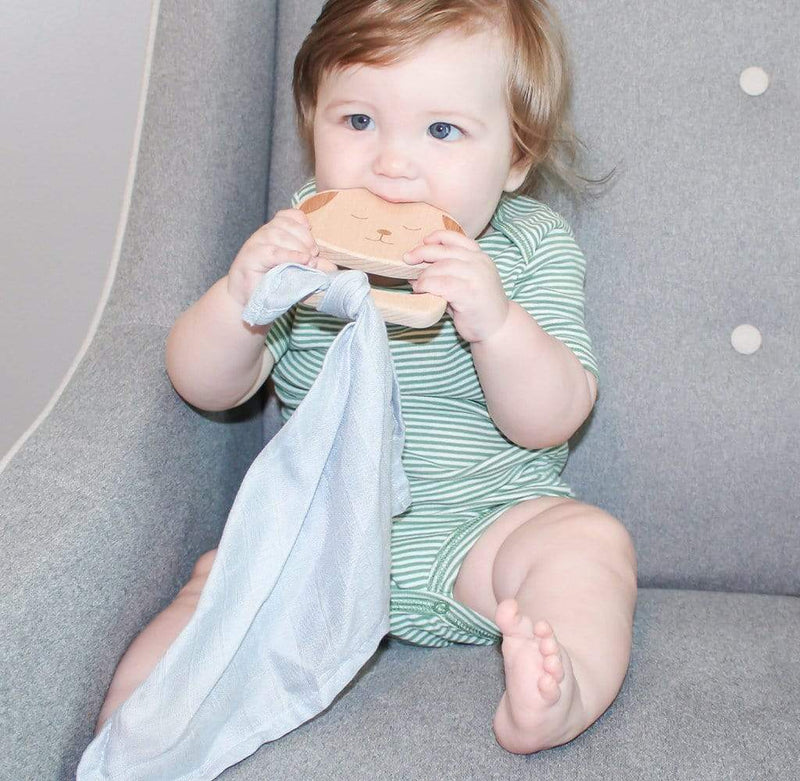 A happy baby sitting on a gray chair, holding the Puppy Wooden Teether & Blue Blankie, joyfully chewing on the teether while wrapped in the cozy blue blankie.