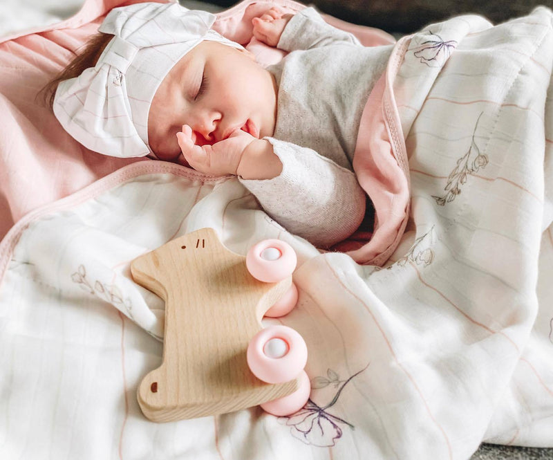 A sleeping baby wrapped in a cozy blanket, holding the Pink Kitten Silicone + Wood Teether with Wheels, illustrating comfort and joy during teething.