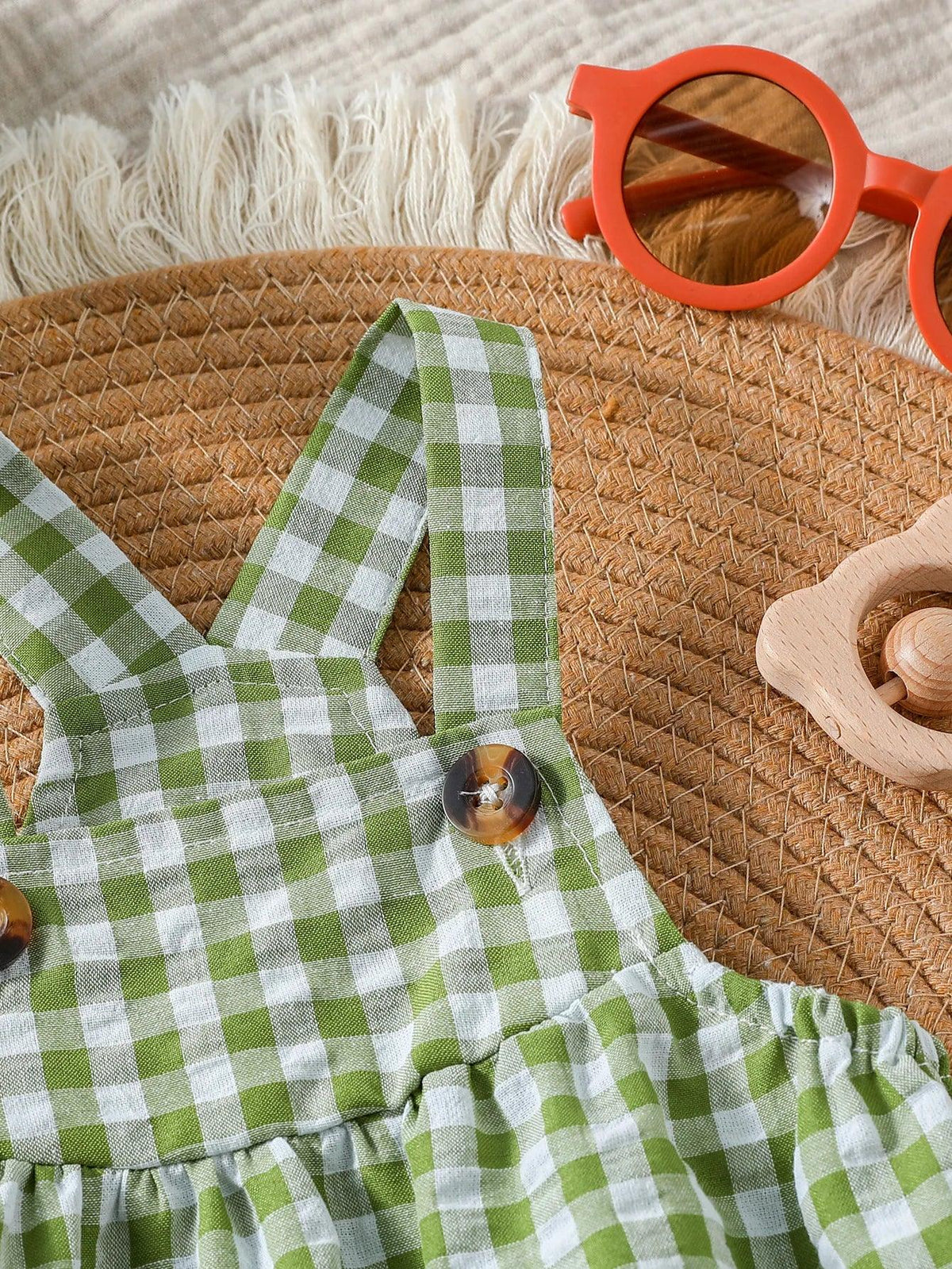 Close-up of green checkered baby romper with buttons on straps lying on woven mat with red sunglasses