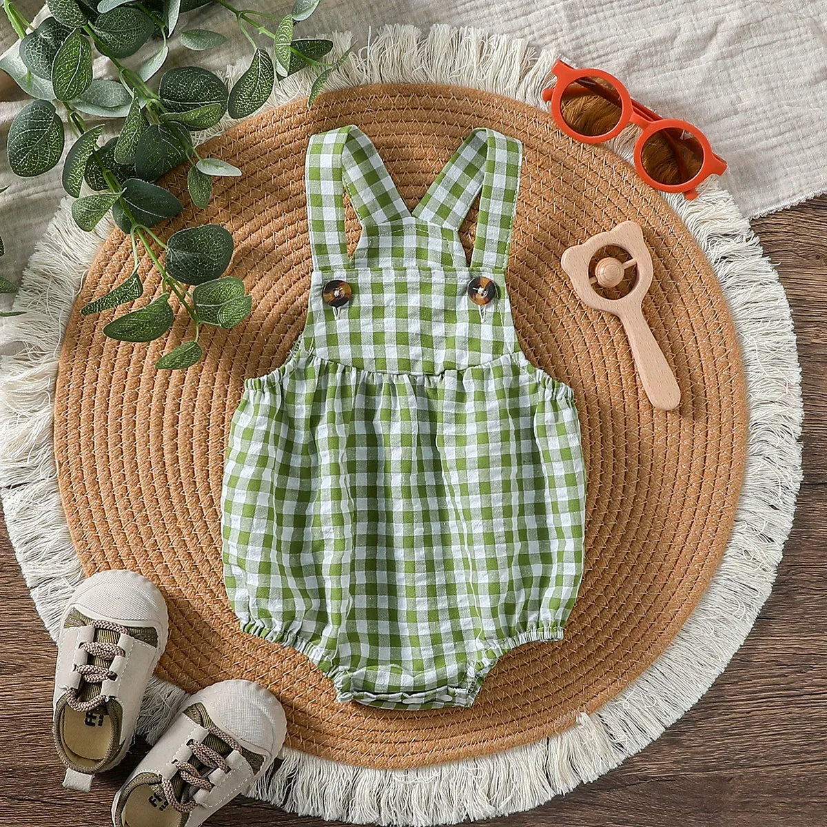 green checkered baby romper laid flat with baby shoes, wooden rattle, and orange sunglasses on round woven mat