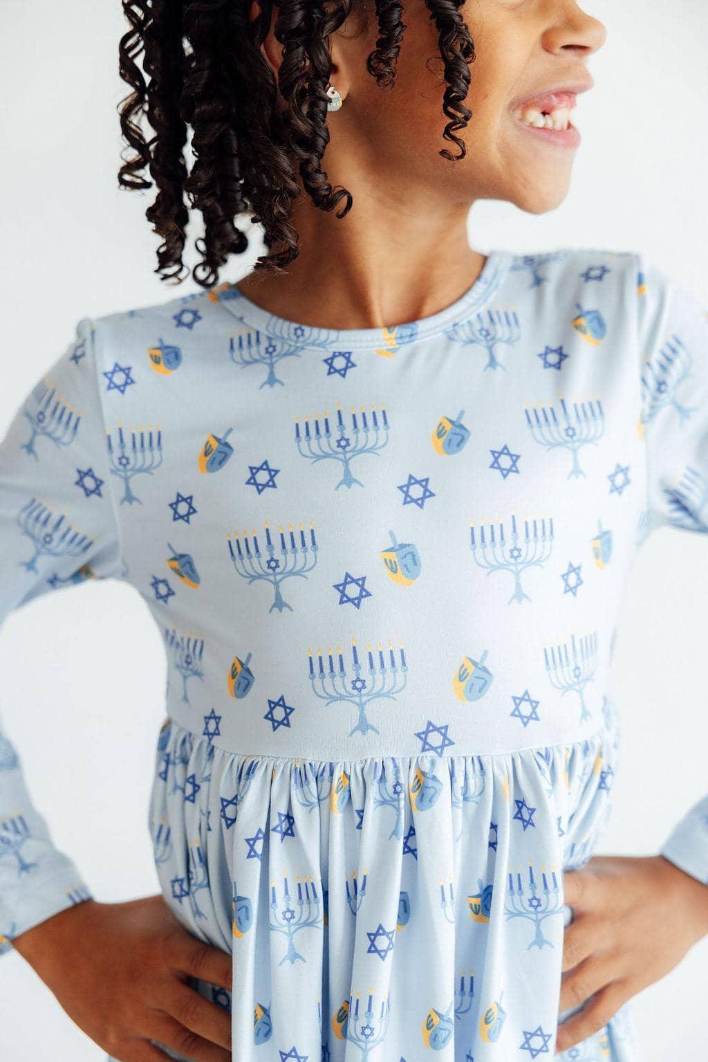 girl wearing light blue Hanukkah dress for girls with menorah and dreidel print smiling with hands on hips