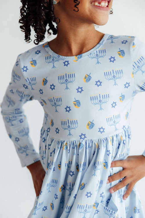 Girl wearing light blue Hanukkah dress for girls with menorah and dreidel patterns posing with hands on hips