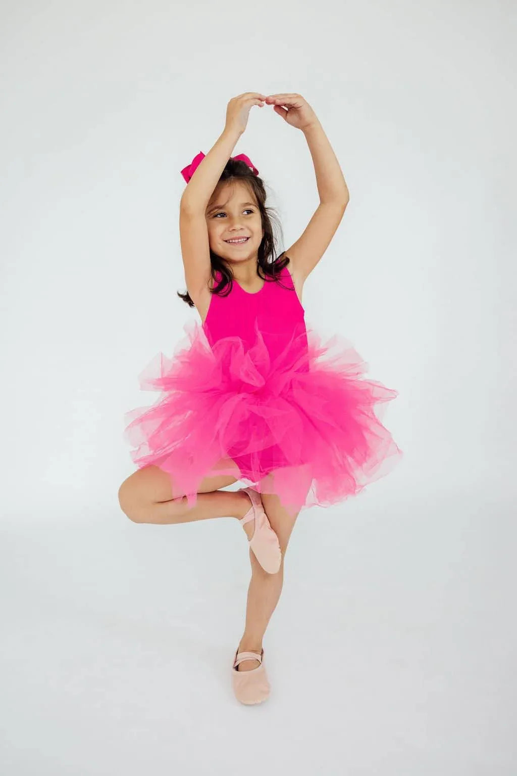young girl wearing hot pink tutu leotard in ballet pose with one leg raised and arms curved above head