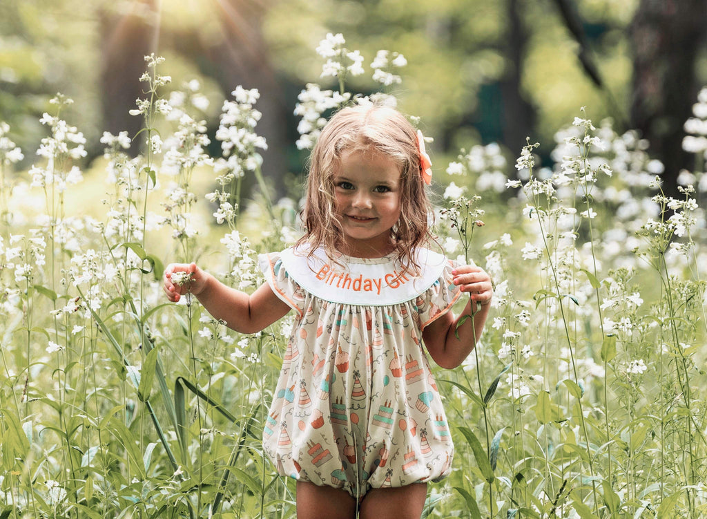 A joyful little girl wearing a 'Cake and Cupcakes' Birthday Girl Smocked Bubble Romper, playing in a field of white flowers, showcasing the vibrant design perfect for any birthday celebration.