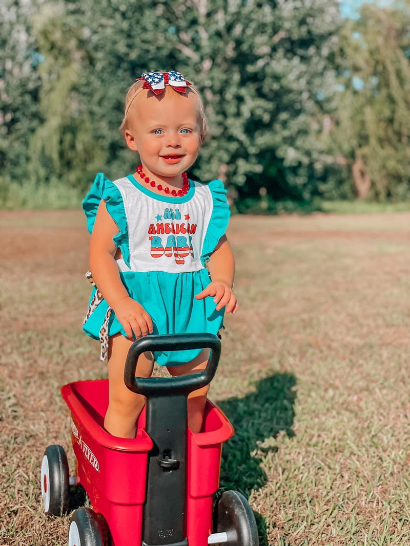 Close-up of a cheerful child in the 'All American Babe' baby bubble romper, standing in a red Radio Flyer wagon against a green backdrop, perfect for summer celebrations.