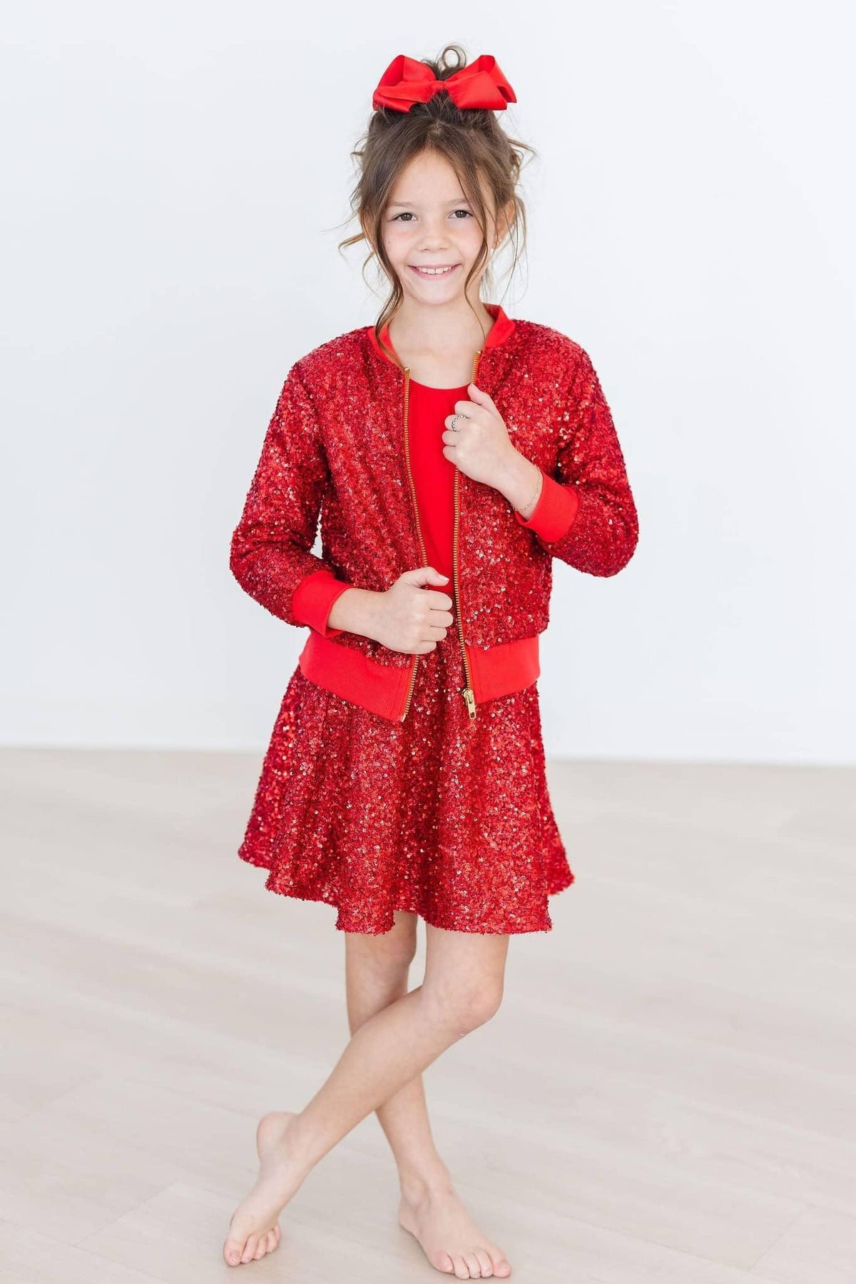 Young girl wearing kids red sequin skort smiling and standing barefoot with red bow in hair