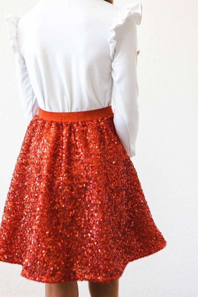 back view of a girl wearing kids red sequin skort with white long sleeve top standing against a white wall