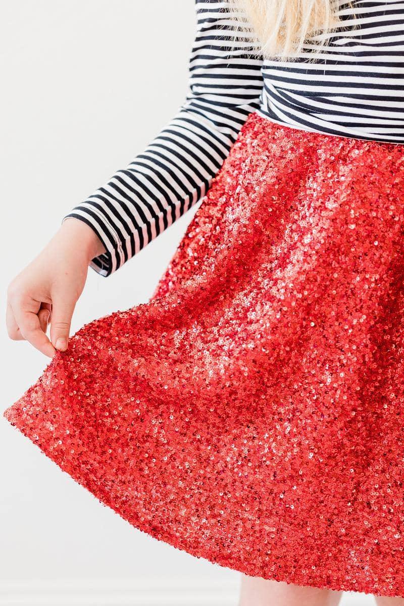 Girl wearing kids red sequin skort holding the hem of the skirt with striped long sleeve top