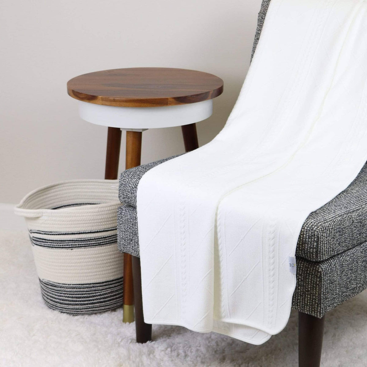 Goosewaddle full size white knit throw draped over gray chair with wooden side table and striped basket