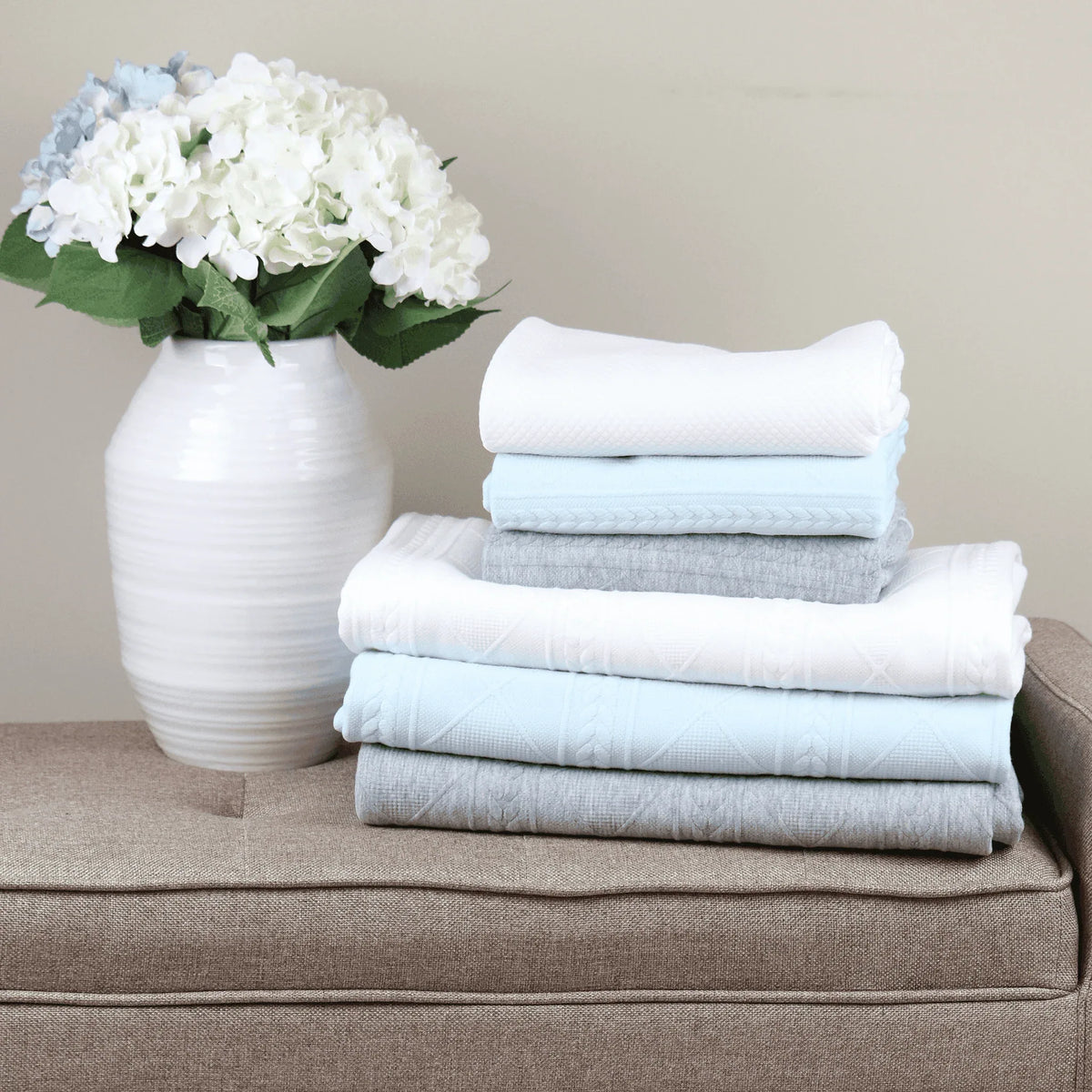 Full size knit throw blankets in white, light blue, and gray stacked next to white vase with flowers