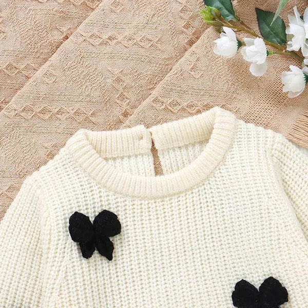 Cream knitted baby romper with bows for infants laid flat on textured beige fabric with white flowers