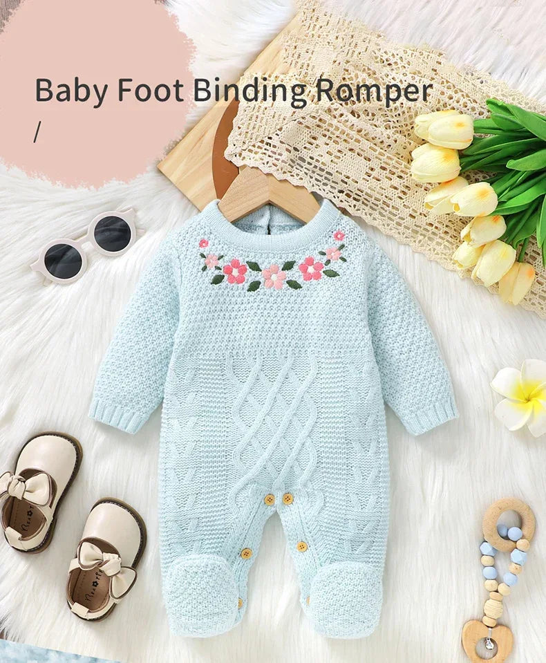 Light blue knitted baby romper with feet for infants laid flat with floral embroidery and surrounded by baby accessories