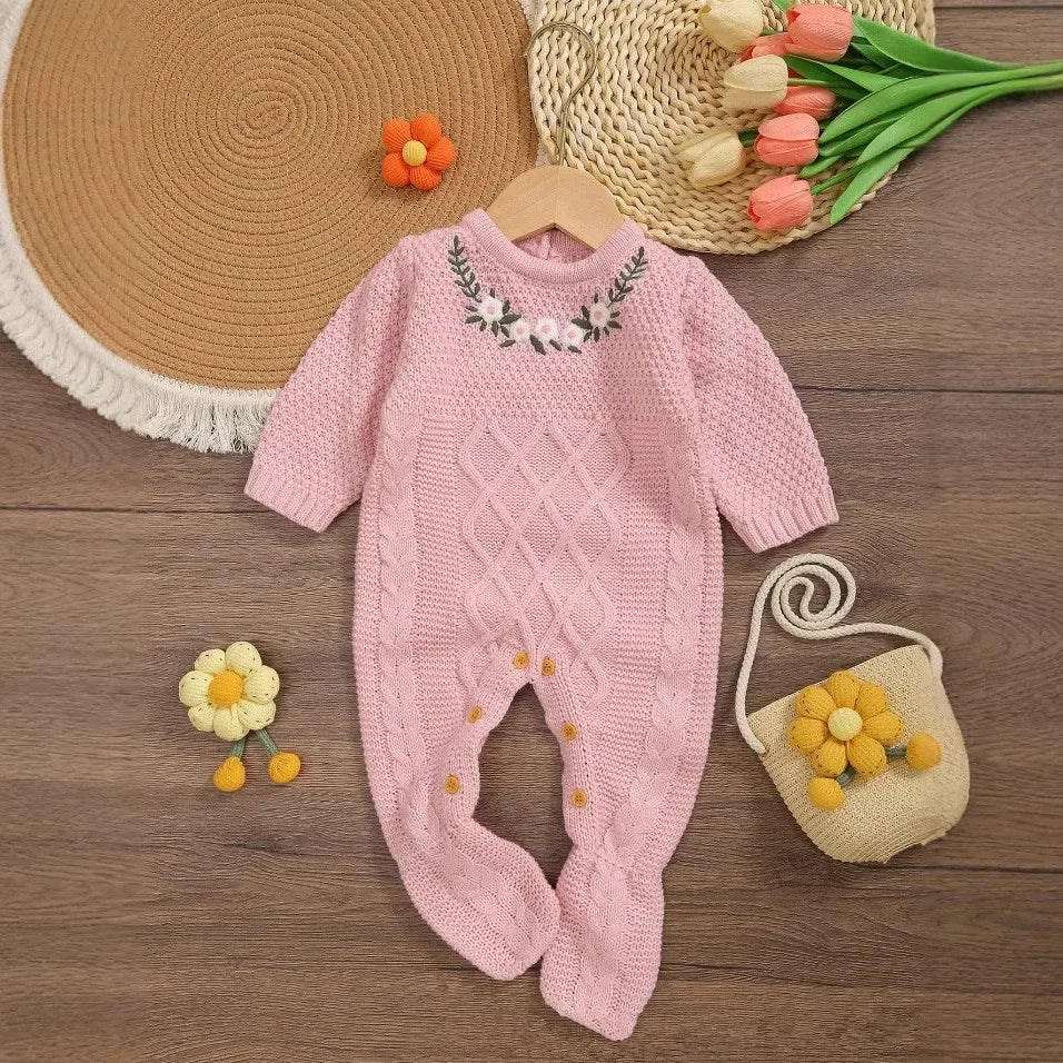 pink knitted baby romper with feet for infants laid flat on wooden floor with floral embroidery on chest