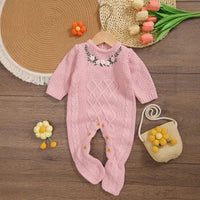 pink knitted baby romper with feet for infants laid flat on wooden floor with floral embroidery on chest