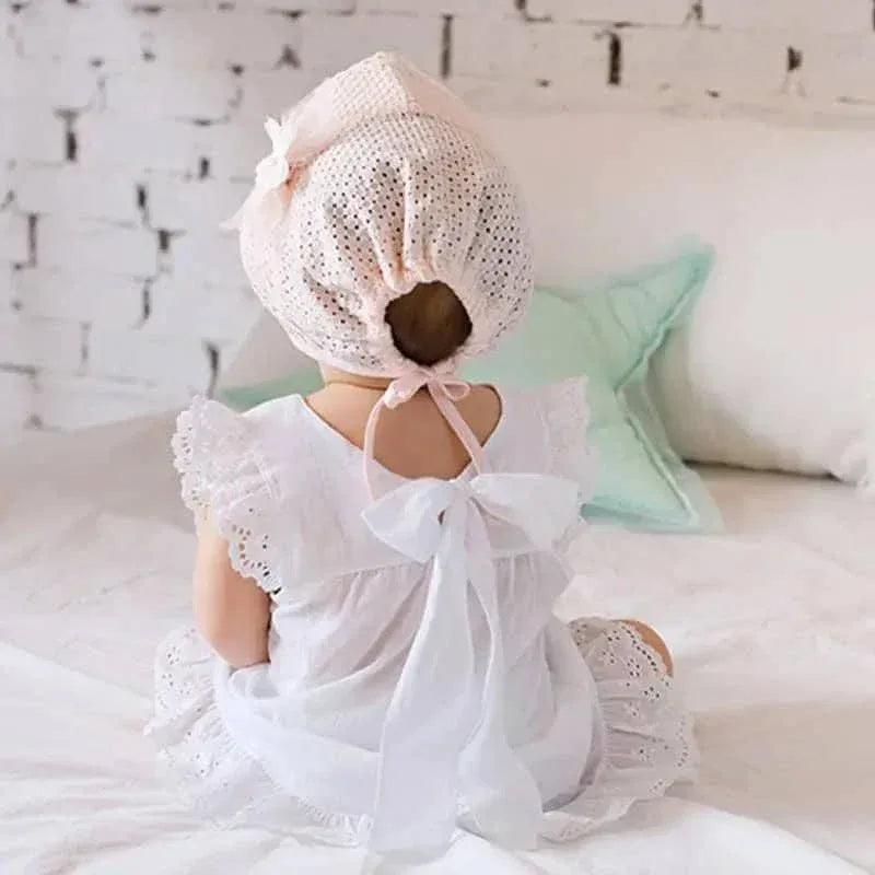 baby wearing white lace baby bonnet and matching white dress sitting on bed facing away