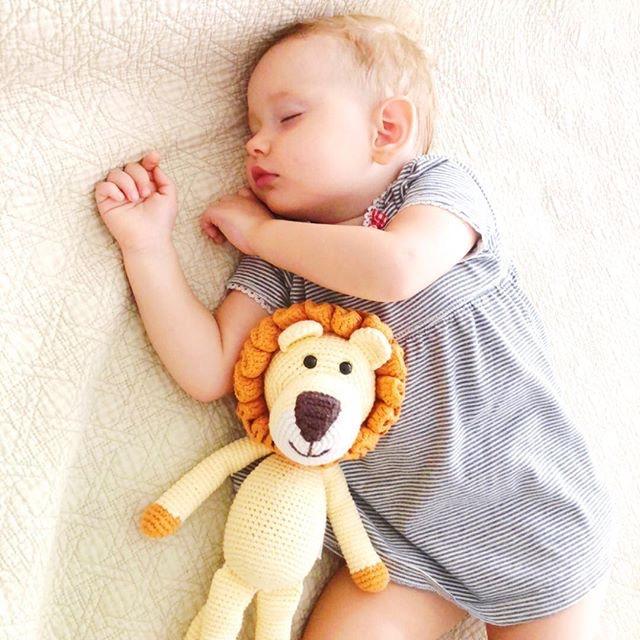 Sleeping baby holds a hand-crocheted Leo lion toy with a golden body and orange mane. A soft organic cotton companion for cuddles.