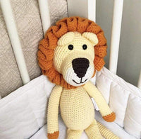 Hand-crocheted lion toy with golden body and orange mane sitting in a white crib. Soft organic cotton stuffed animal for cuddles.
