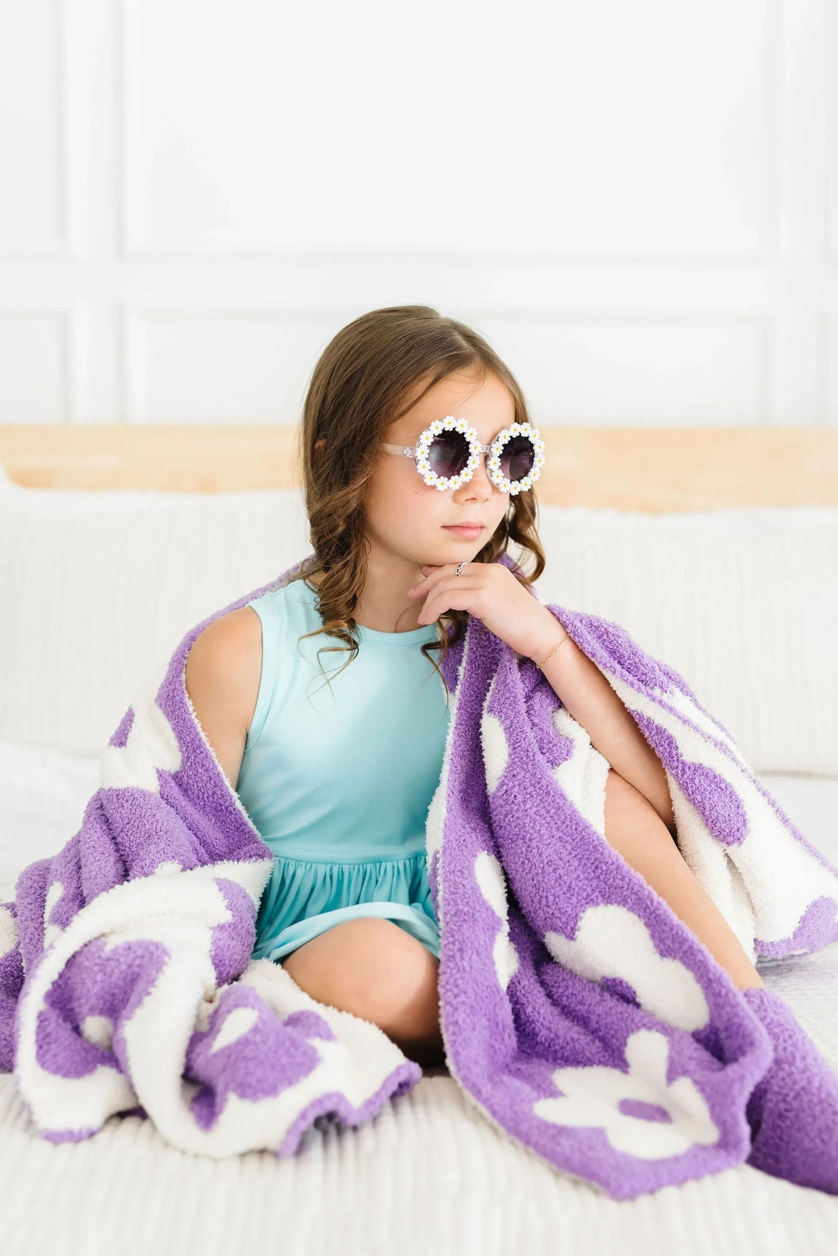 Young girl sitting on couch wrapped in a lilac flower throw blanket for girls from Mila & Rose wearing white flower sunglasses