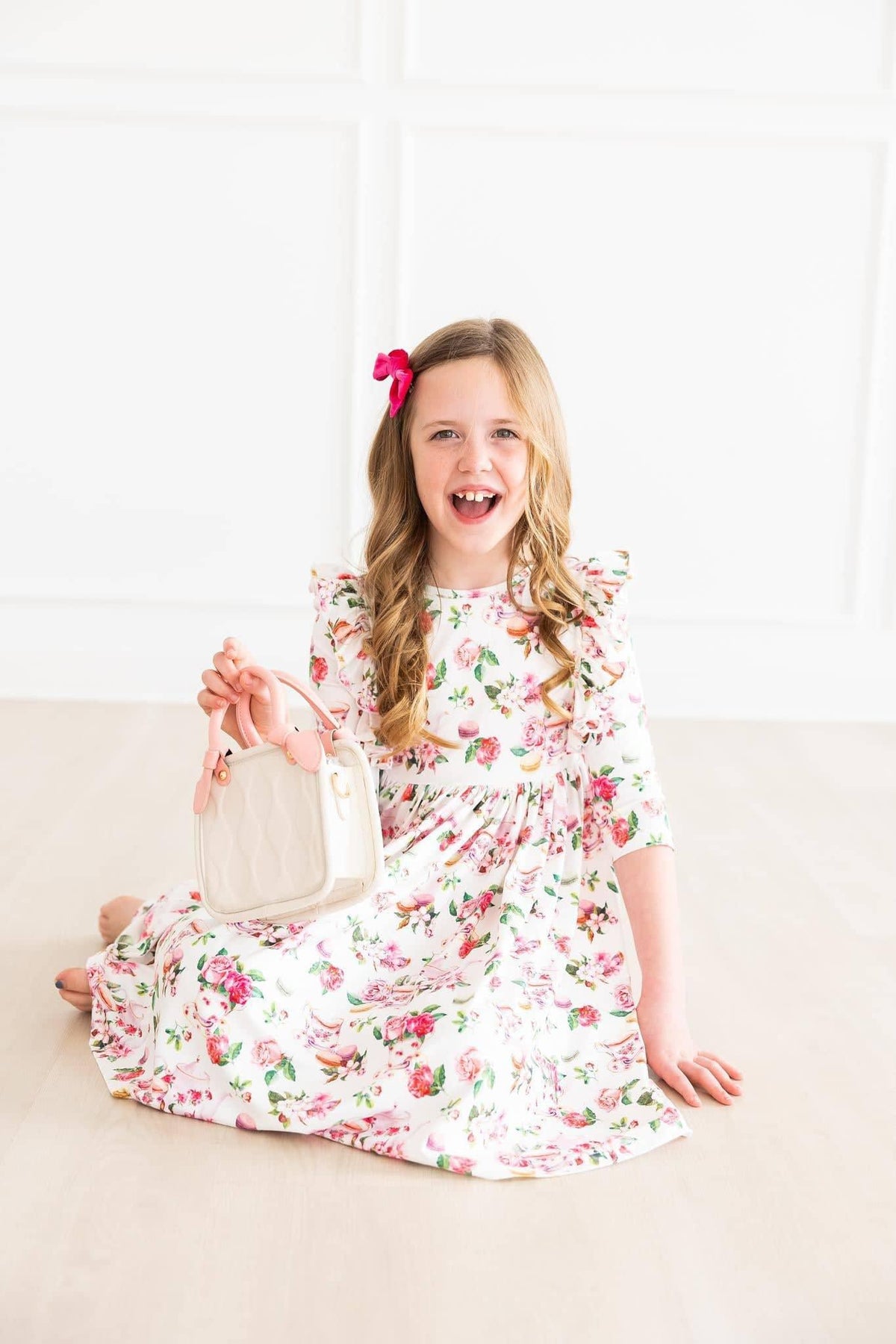 young girl wearing white floral ruffle twirl dress sitting and smiling while holding a white purse