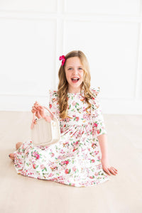 young girl wearing white floral ruffle twirl dress sitting and smiling while holding a white purse