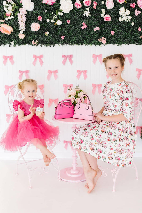 young girl wearing bright pink ruffle twirl dress sitting next to older girl in floral dress holding pink handbags