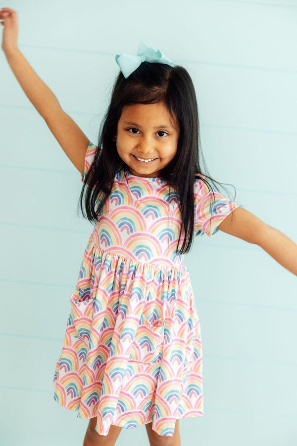 Smiling girl wearing Watercolor Rainbows Short Sleeve Pocket Twirl Dress with pastel rainbow arches and pockets.