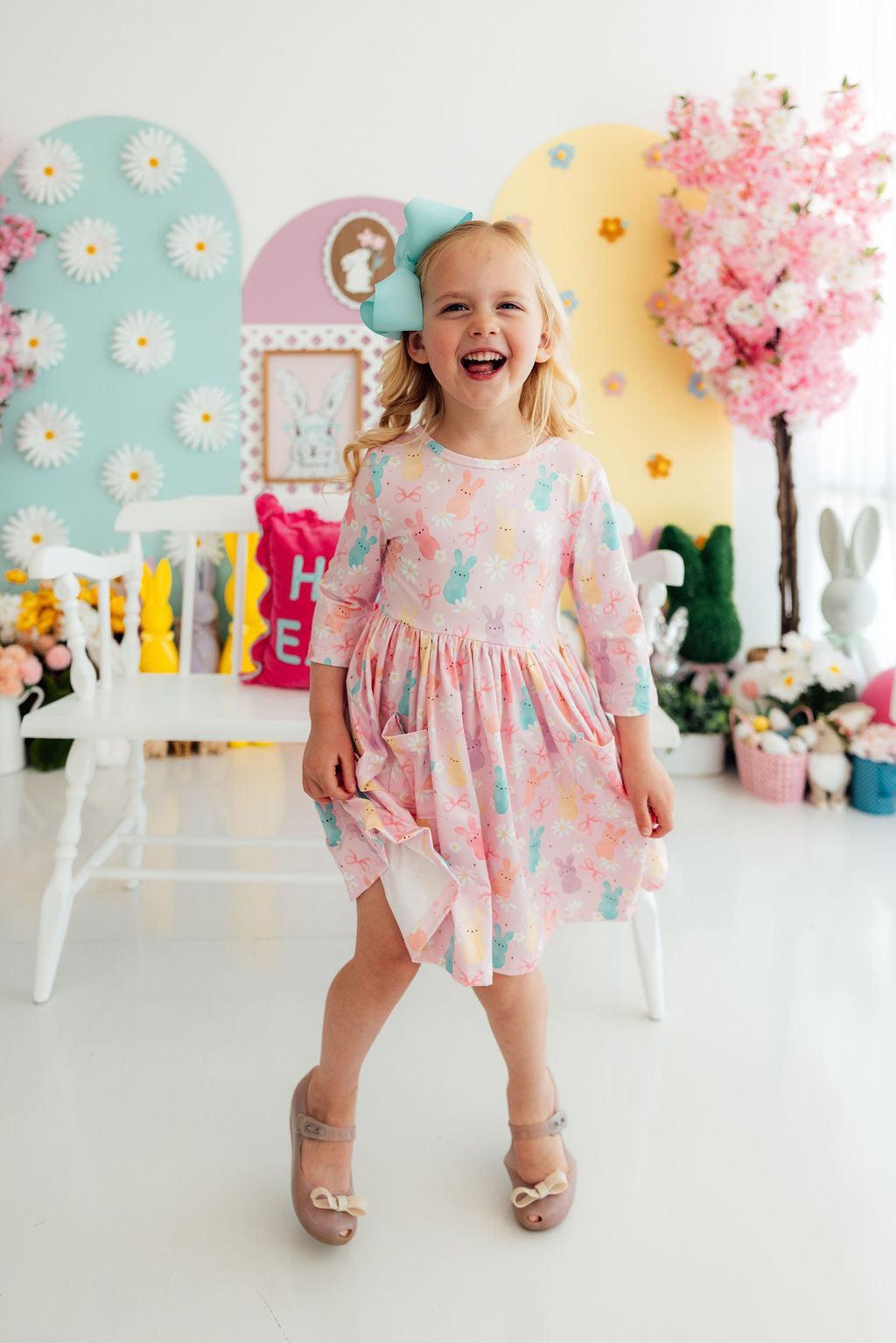 Toddler girl in pastel bunny-print twirl dress with pockets and 3/4 sleeves, smiling in a pastel Easter room.