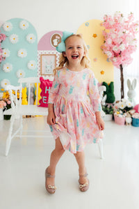 Toddler girl in pastel bunny-print twirl dress with pockets and 3/4 sleeves, smiling in a pastel Easter room.
