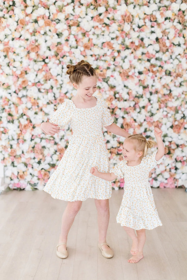 Two girls wearing marigold smocked dress smiling and dancing in front of floral wall