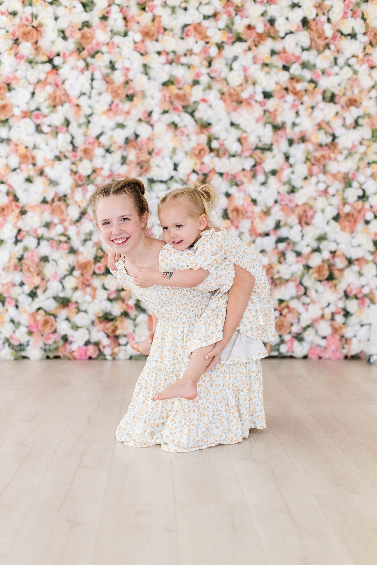 Girls wearing marigold smocked dress, older girl giving younger girl a piggyback in matching floral dresses