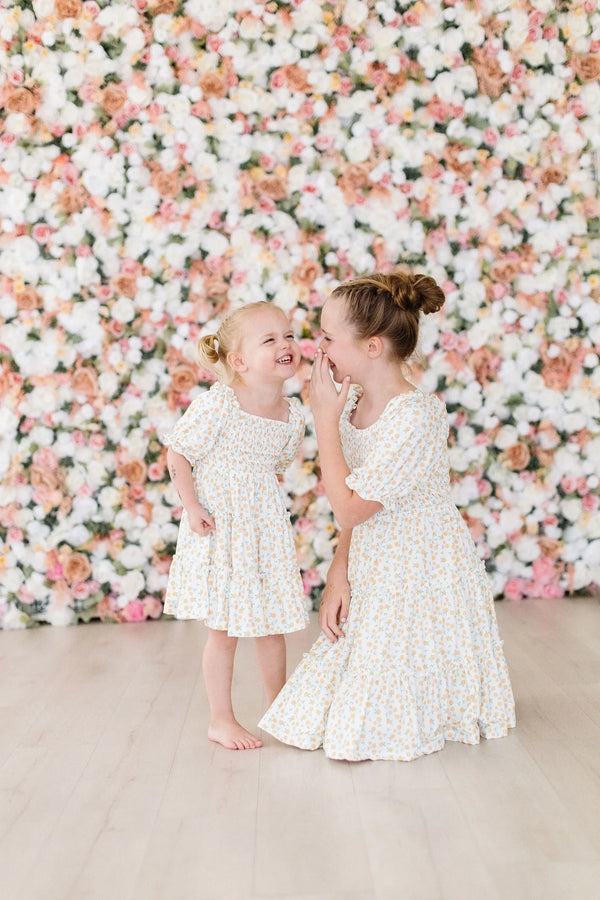 Young girl and toddler wearing marigold smocked dress by Mila Rose smiling and playing in front of floral wall