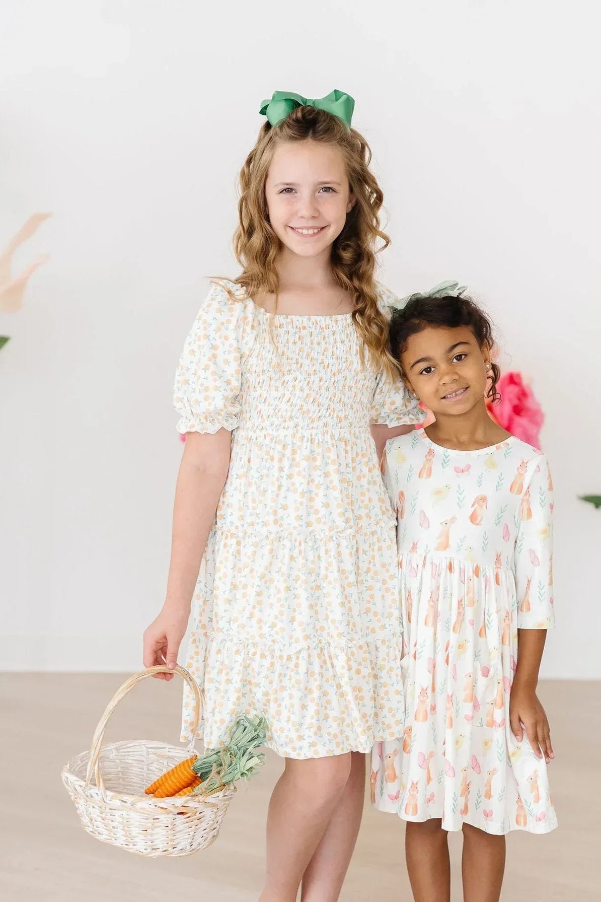 Two girls wearing marigold smocked dress in light colors, one holding a basket with vegetables and smiling