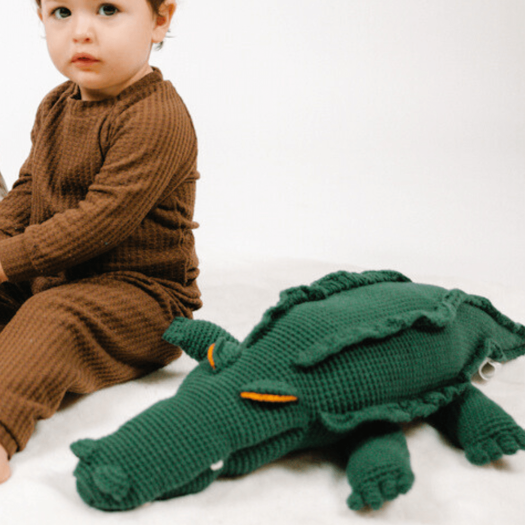 Large green crocheted organic cotton crocodile withridged back and orange eyes, resting beside a child in brown clothing.