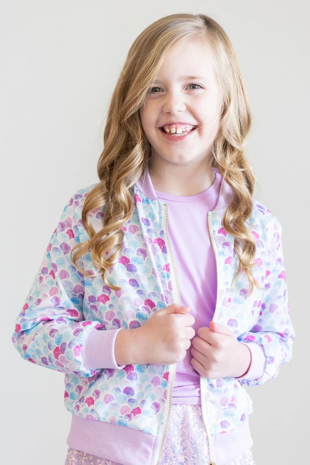 Girl wearing colorful mermaid satin jacket for girls with pastel scales pattern smiling and holding jacket front