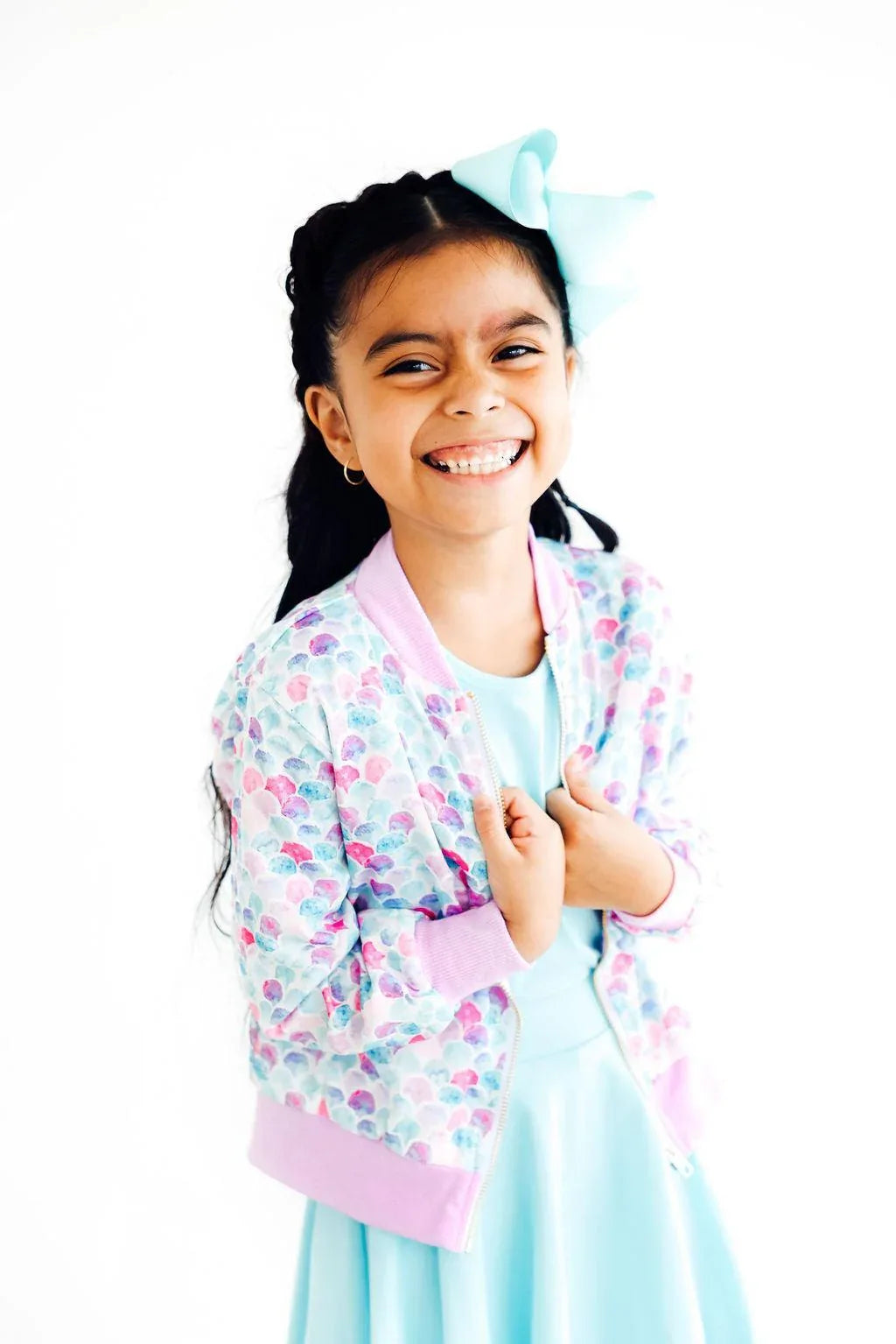 young girl wearing a pink and pastel mermaid satin jacket for girls with a blue dress and bow smiling