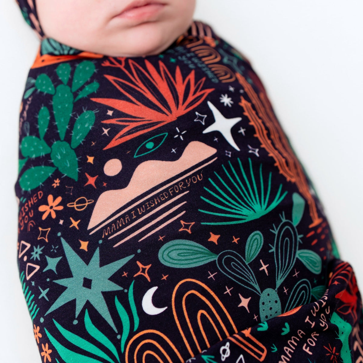 Baby swaddled in The Forever Swaddle Blanket Desert Cactus Midnight featuring green cacti and orange accents.