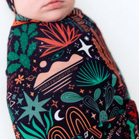 Baby swaddled in The Forever Swaddle Blanket Desert Cactus Midnight featuring green cacti and orange accents.