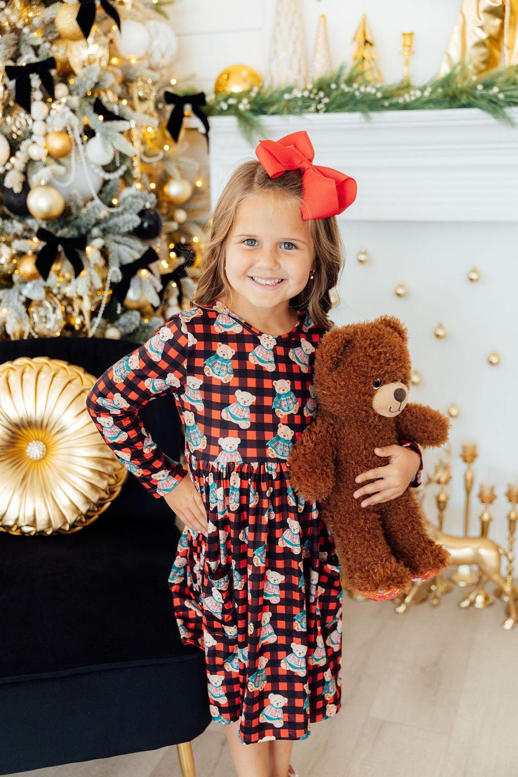 A girl wearing a Christmas Bears Long Sleeve Pocket Twirl Dress poses with a teddy bear, showcasing the festive design and twirl skirt, perfect for holiday celebrations.