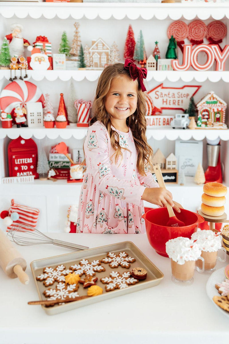 Happy girl preparing holiday treats in a Christmas Cookies Long Sleeve Girls Pocket Twirl Dress, featuring vibrant Christmas-themed patterns. Ideal for festive fun.