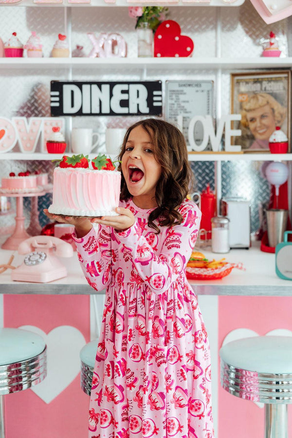 Girl in a pink Vintage Cakes Long Sleeve Pocket Twirl Dress twirls with cake in a retro diner kitchen