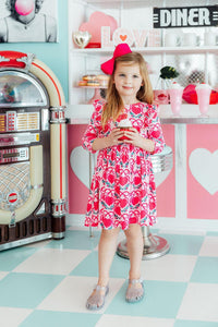 Toddler girl in pink Fluttering Hearts 3/4 sleeve ruffle twirl dress with red bow, holding a cupcake in a retro diner setting.