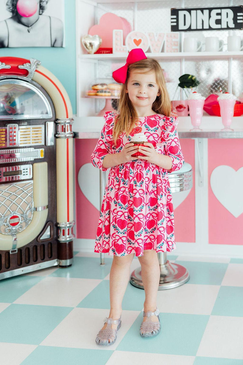 Toddler girl in pink Fluttering Hearts 3/4 sleeve ruffle twirl dress with red bow, holding a cupcake in a retro diner setting.