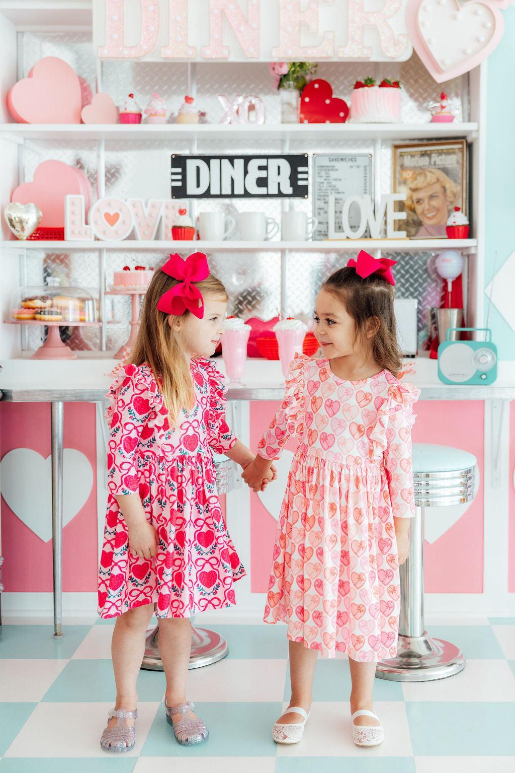 Two toddlers holding hands in a retro diner wearing heart print ruffle twirl dresses with bright pink bows.