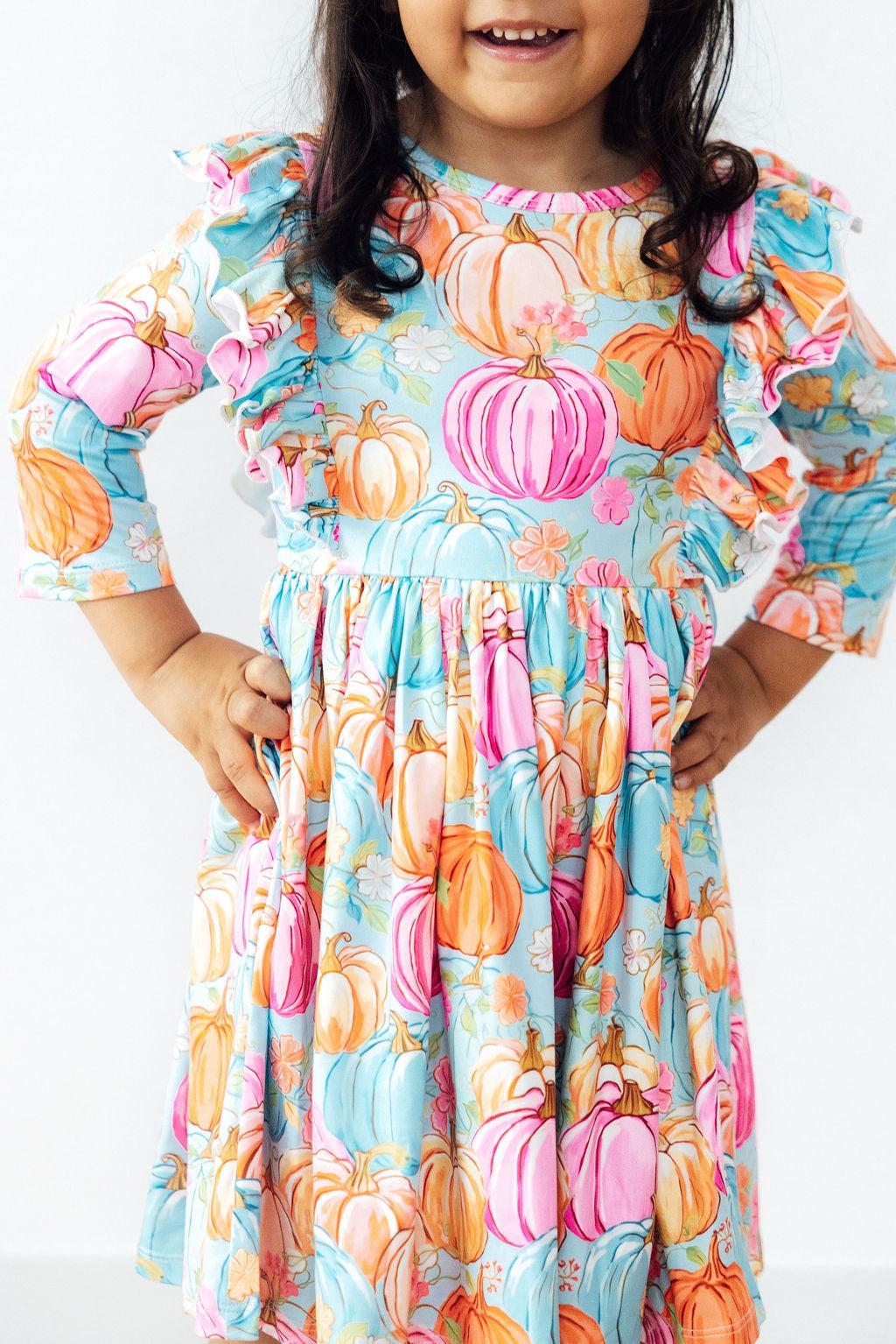 Little girl smiling in a Preppy Pumpkins 3/4 Sleeve Ruffle Twirl Dress by Mila & Rose, showcasing vibrant pumpkin prints in autumn colors.