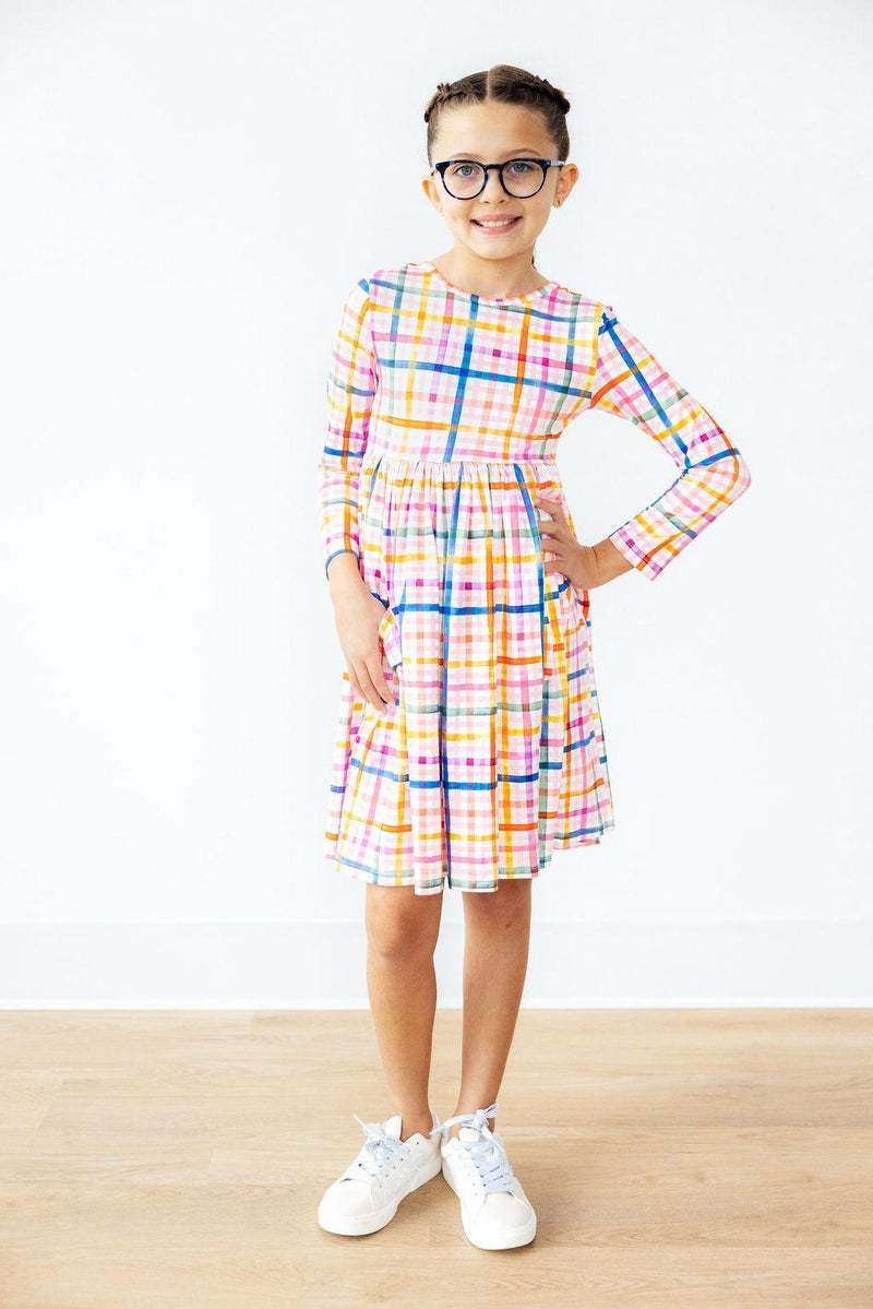 Cheerful girl in Plaid About You L/S Pocket Twirl Dress stands poised in a bright room, highlighting the vibrant plaid pattern and comfortable fit for twirling fun.
