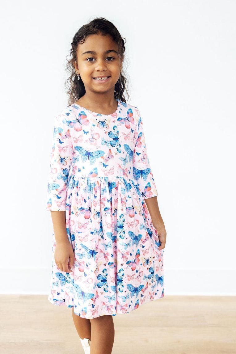 A young girl wearing the Little Flutters 3/4 Sleeve Pocket Twirl Dress with colorful butterfly prints, standing happily with her hands in pockets, perfect for playful outings.