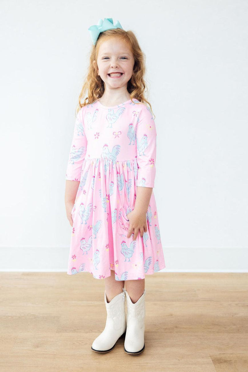A joyful girl smiling in the Little Miss Chickie 3/4 Sleeve Pocket Twirl Dress, showcasing its charming pink design and elegant scoop back.