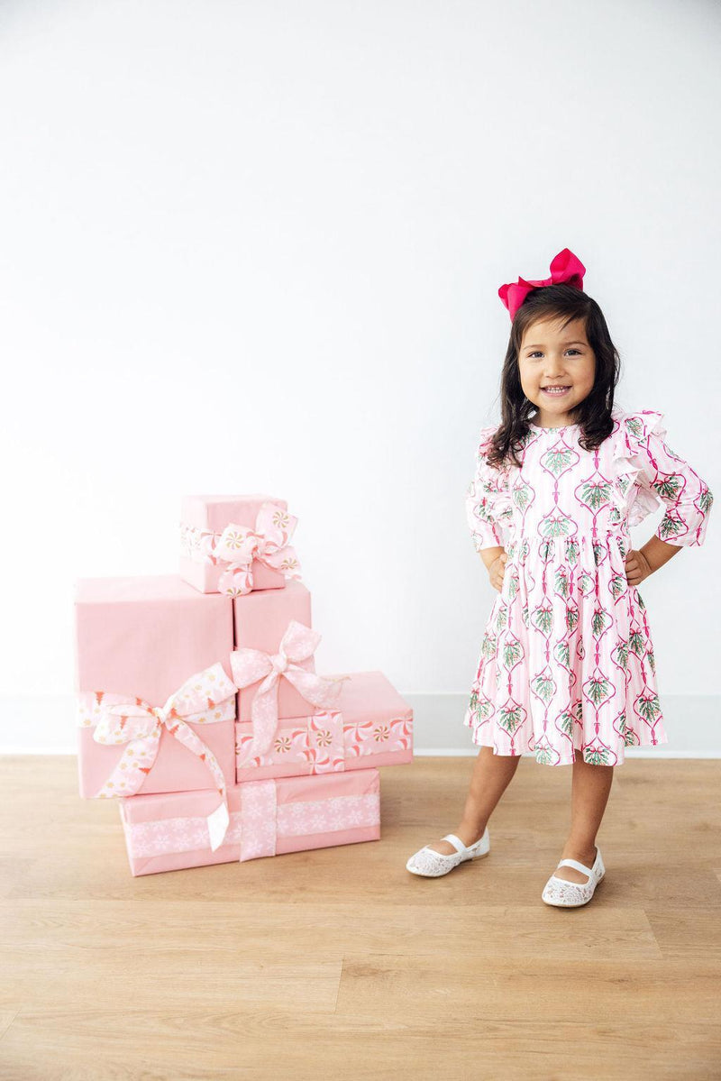 A smiling girl in her Merry Mistletoe 3/4 Sleeve Ruffle Twirl Dress, posing beside stacked pink gift boxes, celebrating the festive spirit.