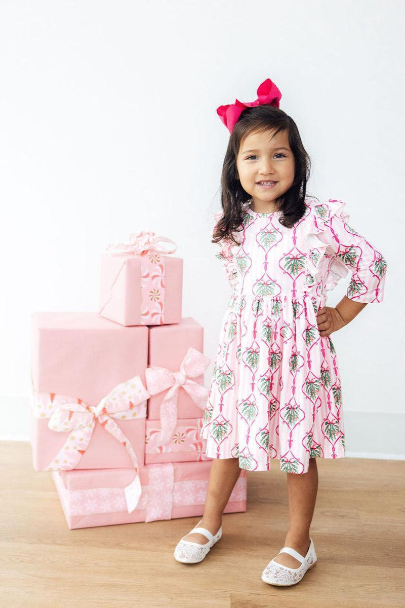 A cheerful little girl in the Merry Mistletoe 3/4 Sleeve Ruffle Twirl Dress, standing confidently by beautifully wrapped pink gifts, ready for Christmas fun.