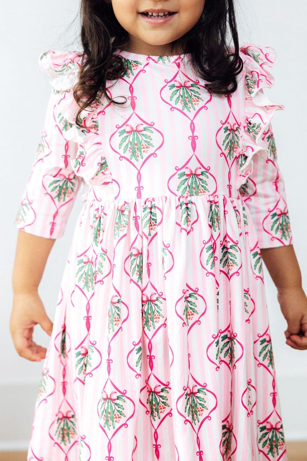 A joyful child wearing the Merry Mistletoe 3/4 Sleeve Ruffle Twirl Dress, showcasing a pink mistletoe pattern with ruffles, perfect for holiday festivities.